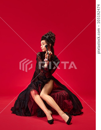 Portrait of beautiful, elegant, tender young girl doing makeup, applying blusher on cheeks against red background Portrait of beautiful, elegant, tender young girl doing makeup, applying blusher on cheeks against red background 103192474