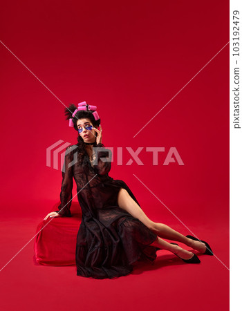 Portrait of young beautiful girl in hair curls and elegant black dress with under eye patches against red background 103192479