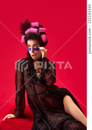 Portrait of young beautiful girl in hair curls and elegant black dress with under eye patches against red background 103192484