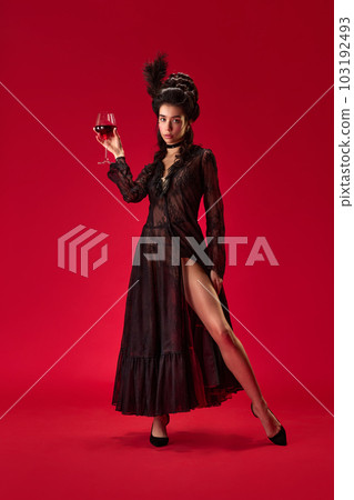 Full-length portrait of beautiful young girl in elegant black dress posing with glass of red wine against red background 103192493