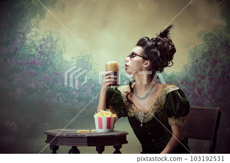 Portrait of beautiful young girl, royal person in vintage dress tasting dark foamy beer with fries against dark canvas background 103192531