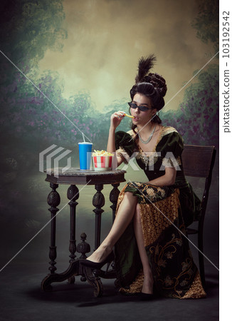 Portrait of young stylish woman, medieval royal person in elegant dress eating fries with soda against dark vintage background 103192542