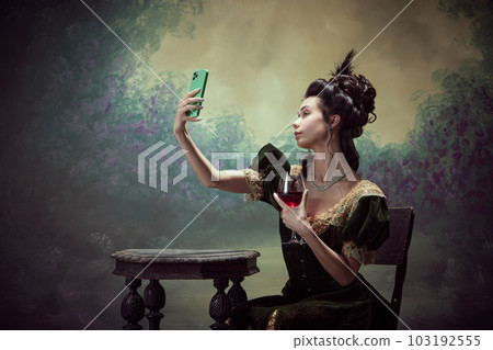 Portrait of young beautiful girl, princess in elegant dress taking selfie with mobile phone and drinking red wine against dark vintage background Portrait of young beautiful girl, princess in elegant dress taking selfie with mobile phone and drinking red wine against dark vintage background 103192555