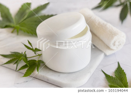 Opened white cream jar with a lid near cannabis leaves. Cosmetic Mockup Opened white cream jar with a lid near cannabis leaves. Cosmetic Mockup 103194044