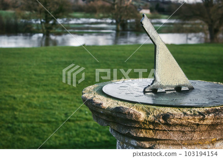 An old metal sundial set on a stone plinth or pedestal on a grassy lawn with a lake in the background 103194154
