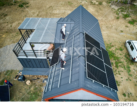 Men workers prepearing for mounting photovoltaic solar modules on roof of house. Electricians in helmets installing solar panel system outdoors. Concept of alternative and renewable energy. 103194716