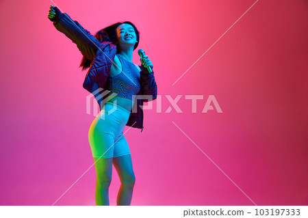Portrait of happy, smiling, young girl singing in microphone against pink studio background in neon light. Leisure 103197333