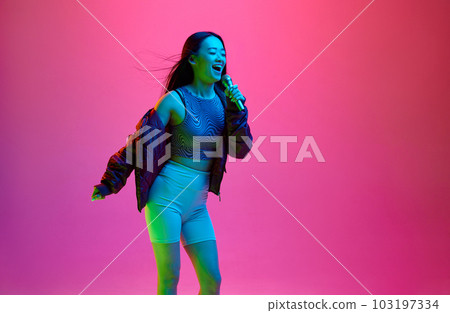 Portrait of happy, smiling, young girl singing in microphone against pink studio background in neon light. Karaoke fun 103197334