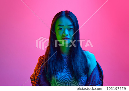 Portrait of young korean girl looking at camera with little smile, posing against pink studio background in neon light 103197359