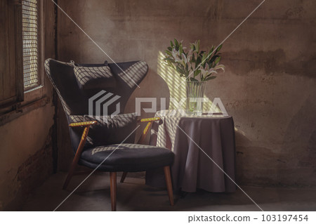 A Wooden Upholstered Chair and the Bouquet of Flowers in a Glass Vase on Table in the Room with Bare Cement Wall. A Wooden Upholstered Chair and the Bouquet of Flowers in a Glass Vase on Table in the Room with Bare Cement Wall. 103197454