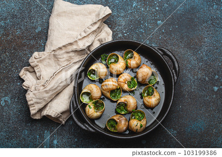 Escargots de Bourgogne Cooked Snails with Garlic Butter and Parsley in black cast iron pan on rustic stone background from above, traditional French Delicacy . 103199386
