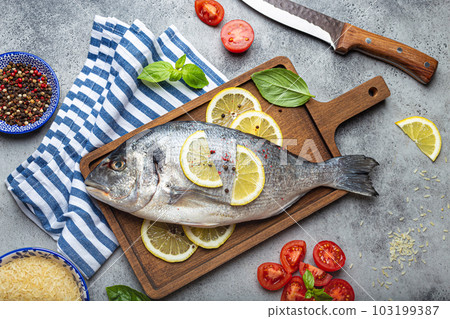 Raw fish dorado with ingredients lemon, fresh basil, cut cherry tomatoes, uncooked rice on wooden cutting board with knife on rustic stone background top view, cooking healthy fish dorado 103199387