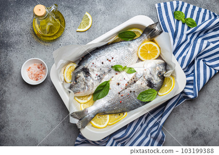 Raw fish dorado in casserole dish with ingredients lemon, fresh basil, bottle of olive oil on wooden cutting board with knife on rustic stone background top view, cooking healthy fish dorado 103199388