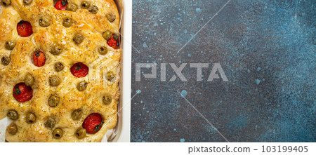 Overhead of Traditional Italian Homemade Flat Bread Focaccia with Green Olives, Olive Oil, Cherry Tomatoes and Rosemary in Baking Tray on Rustic Dark Blue Stone Background, Copy Space 103199405