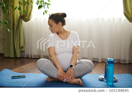 Charming pregnant woman practicing pregnancy yoga at home , sitting on a fitness mat in lotus posture and using smartphone, watching online class with breathing exercises and prenatal stretching. 103199695