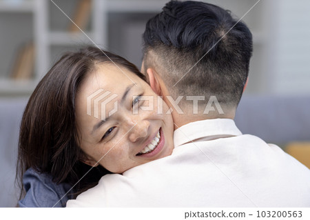 Close-up photo. Young Asian couple, family hugging together. Happy face of young woman hugging and supporting her husband, meets smiling at home. Close-up photo. Young Asian couple, family hugging together. Happy face of young woman hugging and supporting her husband, meets smiling at home. 103200563