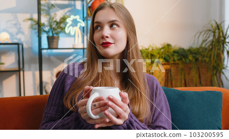 Pretty smiling young woman drinking a cup of coffee or herbal tea sitting at home couch, break time 103200595