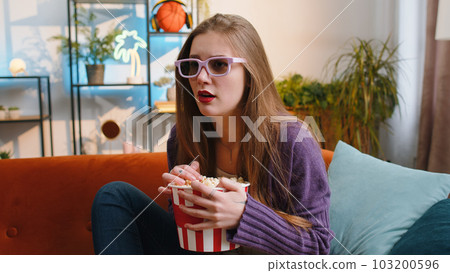 Pretty woman sits on sofa eating popcorn, watching interesting TV serial, sport game online at home 103200596