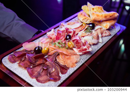 The waiter holds a wooden plate with various sliced dried meats delicacies The waiter holds a wooden plate with various sliced dried meats delicacies 103200944