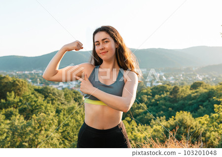 Young sporty woman shows pumped up arm muscles during training in the Park. The concept of yoga, sports and meditation. In the background view of the settlement and the mountains. Copy space Young sporty woman shows pumped up arm muscles during training in the Park. The concept of yoga, sports and meditation. In the background view of the settlement and the mountains. Copy space 103201048