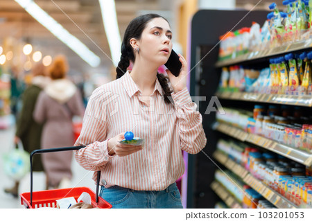Portrait of young pretty woman holds cart and products and calls with cellphone. Shelves with food in background. Concept of shopping in grocery store Portrait of young pretty woman holds cart and products and calls with cellphone. Shelves with food in background. Concept of shopping in grocery store 103201053