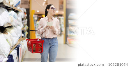 Web banner of shopping with mock up. Young caucasian woman wearing glasses holds cart and thoughtfully choises pillow and bedding. Concept of supermarket purchase 103201054