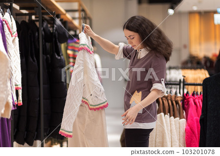 Young Caucasian woman chooses sweater in store. In background there are d hangers with clothes. Concept of shopping and purchase Young Caucasian woman chooses sweater in store. In background there are d hangers with clothes. Concept of shopping and purchase 103201078