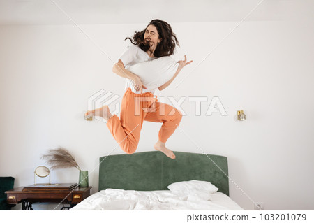 Happy young Caucasian woman funny jumping on bed and play on air guitar. Concept of happiness and fun 103201079