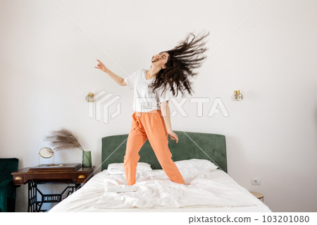 Happy young Caucasian woman dancing on bed in bedroom. Dark hair fly away. Freedom and relax at home. Happy young Caucasian woman dancing on bed in bedroom. Dark hair fly away. Freedom and relax at home. 103201080