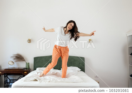 Happy young Caucasian woman enjoying and dancing on bed in bedroom. Enjoying winning. Freedom and relax at home Happy young Caucasian woman enjoying and dancing on bed in bedroom. Enjoying winning. Freedom and relax at home 103201081