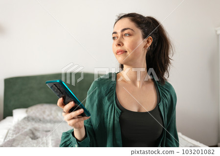 Portrait of young Caucasian woman using smartphone sitting on bed. Concept of online casino game and social media. Portrait of young Caucasian woman using smartphone sitting on bed. Concept of online casino game and social media. 103201082