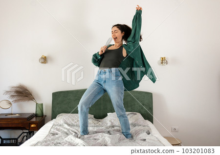 Young Caucasian happy woman is singing and dancing on bed. Funny enjoy in bedroom. Concept of dance and vocals learning Young Caucasian happy woman is singing and dancing on bed. Funny enjoy in bedroom. Concept of dance and vocals learning 103201083
