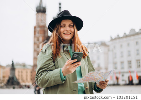 Beautiful stylish woman in hat walking on Market Square in Krakow on autumn day and holding mobile phone and map. Phone Communication. Urban lifestyle concept. Check social networks, booking hotel 103201151