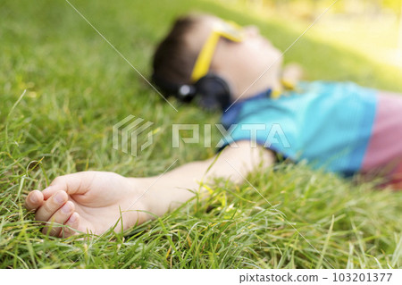 Happy boy in sunglasses lying on the grass and listening to music in headphones Happy boy in sunglasses lying on the grass and listening to music in headphones 103201377