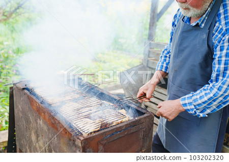 an elderly man with a beard in work clothes cooks meat steaks on the grill  103202320