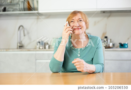 Elderly positive woman stands at home in kitchen and talks nicely on phone with friend Elderly positive woman stands at home in kitchen and talks nicely on phone with friend 103206530