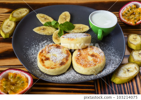 Syrniki or cheese pancakes with cream sauce and bananas on a plate on wooden table close up Syrniki or cheese pancakes with cream sauce and bananas on a plate on wooden table close up 103214075