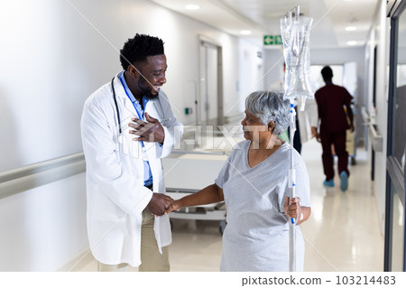 Biracial senior female patient with drip shaking hand of happy diverse male doctor in corridor 103214483