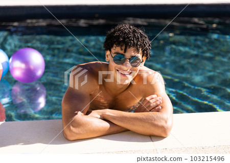 Portrait of happy fit shirtless biracial man in sunglasses in swimming pool with arms crossed in sun 103215496