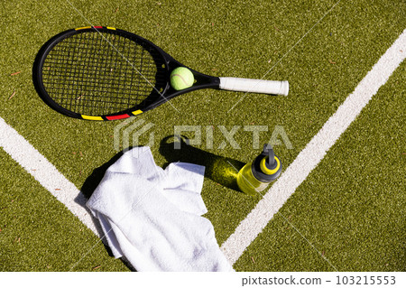 Tennis ball, racket, towel and water bottle lying on sunny outdoor grass tennis court 103215553