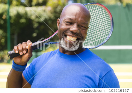 Portrait of happy senior african american man with tennis racket at sunny grass court 103216137