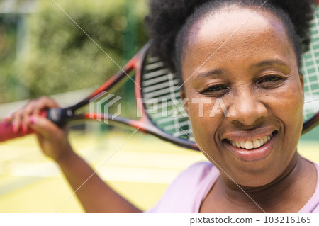 Portrait of senior african american woman with tennis racket smiling on sunny grass court 103216165