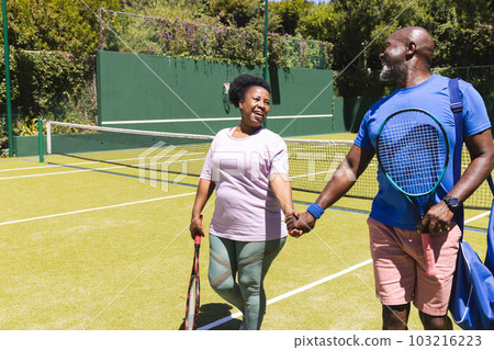 Happy senior african american couple with rackets walking holding hands on sunny grass tennis court 103216223