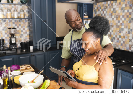 Happy senior african american couple preparing meal together, using tablet in kitchen 103216422