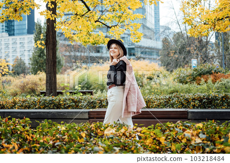 Fashion portrait of stylish young woman in hat walking in the autumn city. Happy gen z woman walking in the city with yellow autumn foliage 103218484