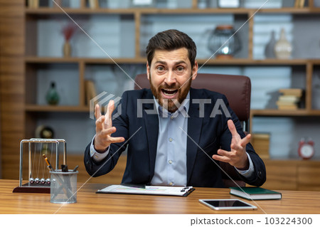Angry and screaming businessman inside office talking on video call, boss shouting in online meeting with colleagues and subordinates, dissatisfied with work of colleagues and partners. 103224300