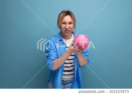 blond neat cheerful girl with a grimace holds a piggy bank on a blue background blond neat cheerful girl with a grimace holds a piggy bank on a blue background 103225973