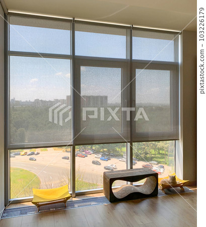 Roller blinds on the windows in the interior. Automatic solar shades on full height windows. Motorized sunscreen blinds with screen fabric. Ginger cat lies on a couch near the window with curtains. Roller blinds on the windows in the interior. Automatic solar shades on full height windows. Motorized sunscreen blinds with screen fabric. Ginger cat lies on a couch near the window with curtains. 103226179