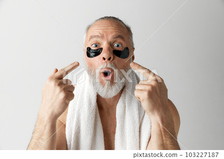 Portrait of handsome mature man with towel, shirtless posing with under-eye patches against grey background. Reducing tired look Portrait of handsome mature man with towel, shirtless posing with under-eye patches against grey background. Reducing tired look 103227187