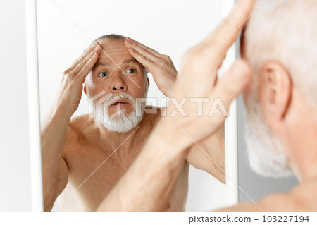 Portrait of mature shirtless man with beard and gray hair, looking in mirror at wrinkled forehead against grey background 103227194